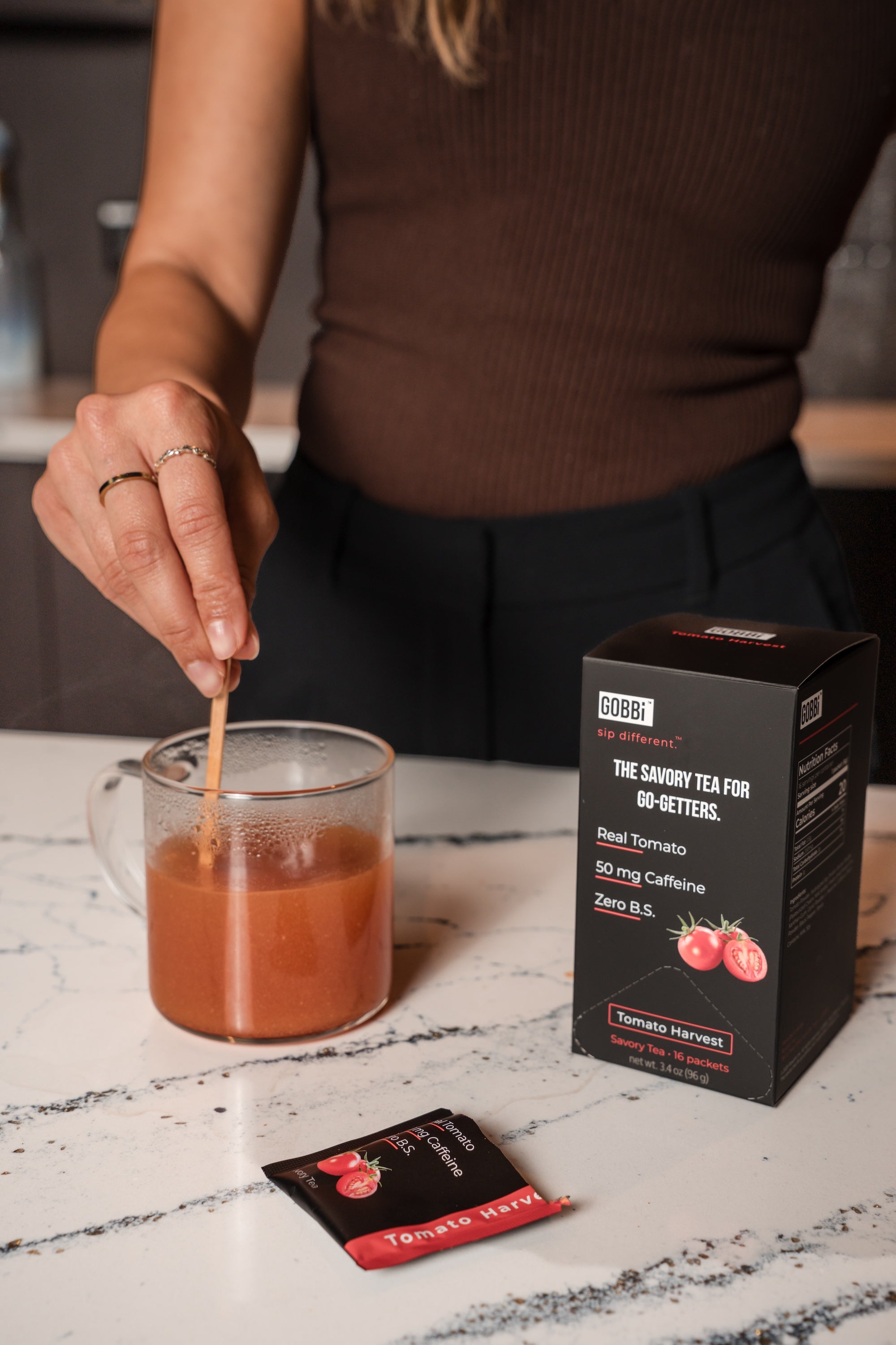 Person stirring a mug of tea with a Gobbi tomato harvest Tea box and packets on a marble surface.