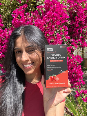 Roma, the founder of Gobbi, holding a Gobbi Savory Energy Drink package in front of pink flowers